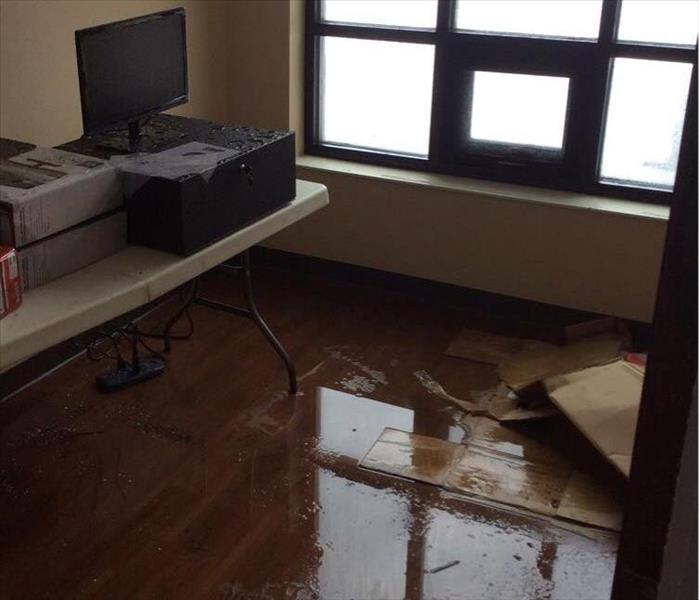 A desk with a computer and boxes. Water on a hardwood floor and a window in the background